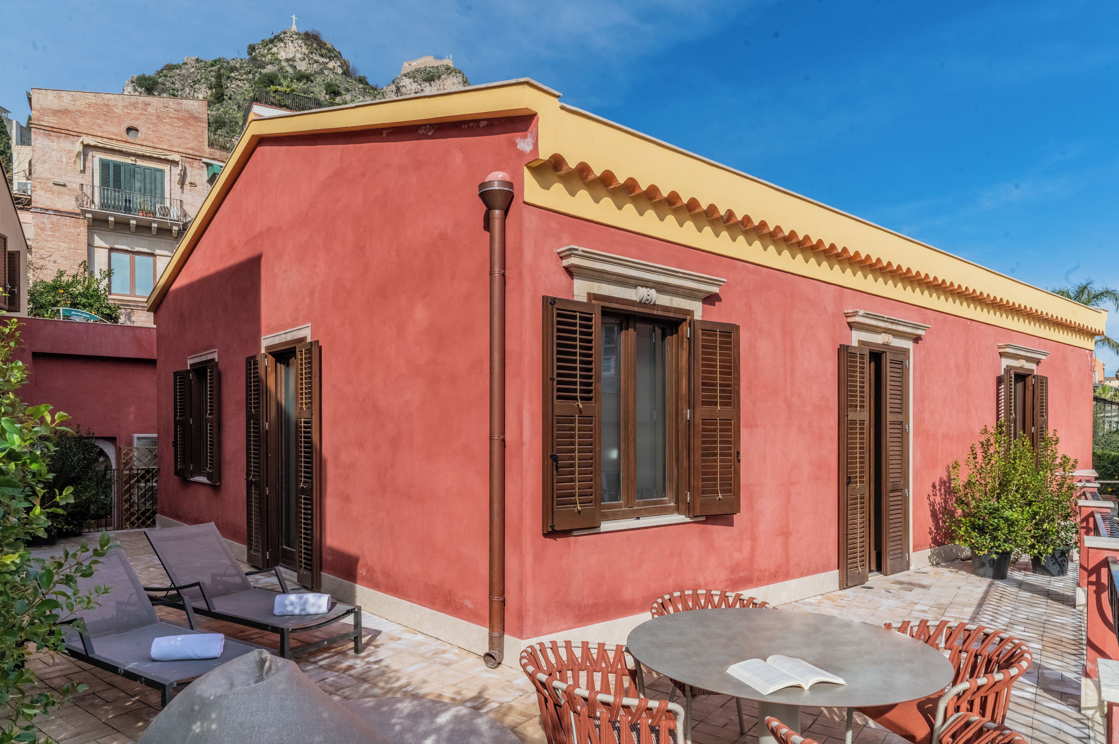 Un edificio rojo con techo amarillo, persianas marrones y un patio con baldosas y mobiliario exterior.