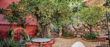An outdoor patio with a brick floor, red and white walls, and orange trees. There are round tables and chairs, and a sofa.