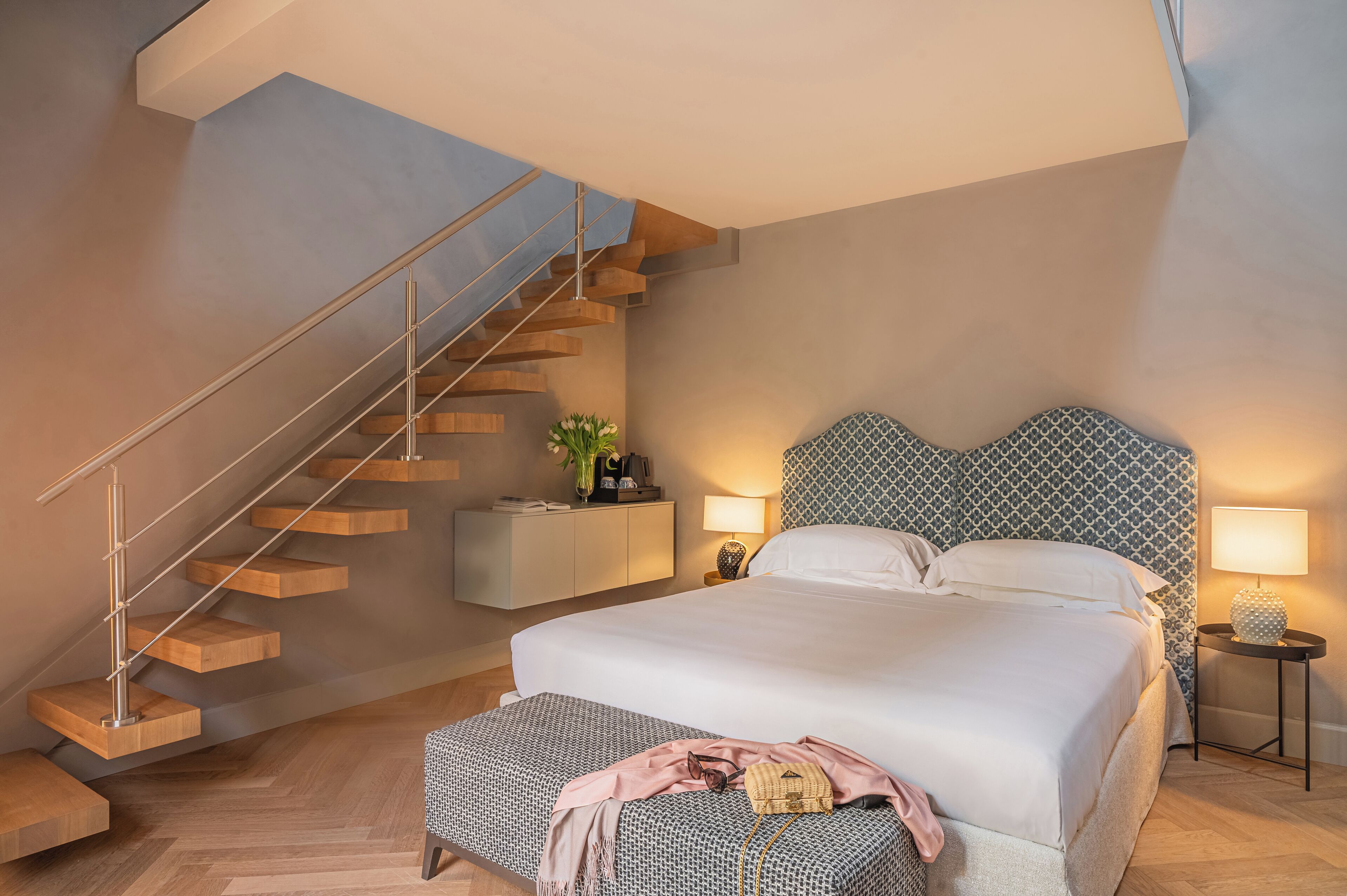 Un dormitorio de diseño moderno, con una escalera de madera, cama con cabecera estampada, lámparas de noche y una mesita de noche con una planta.
