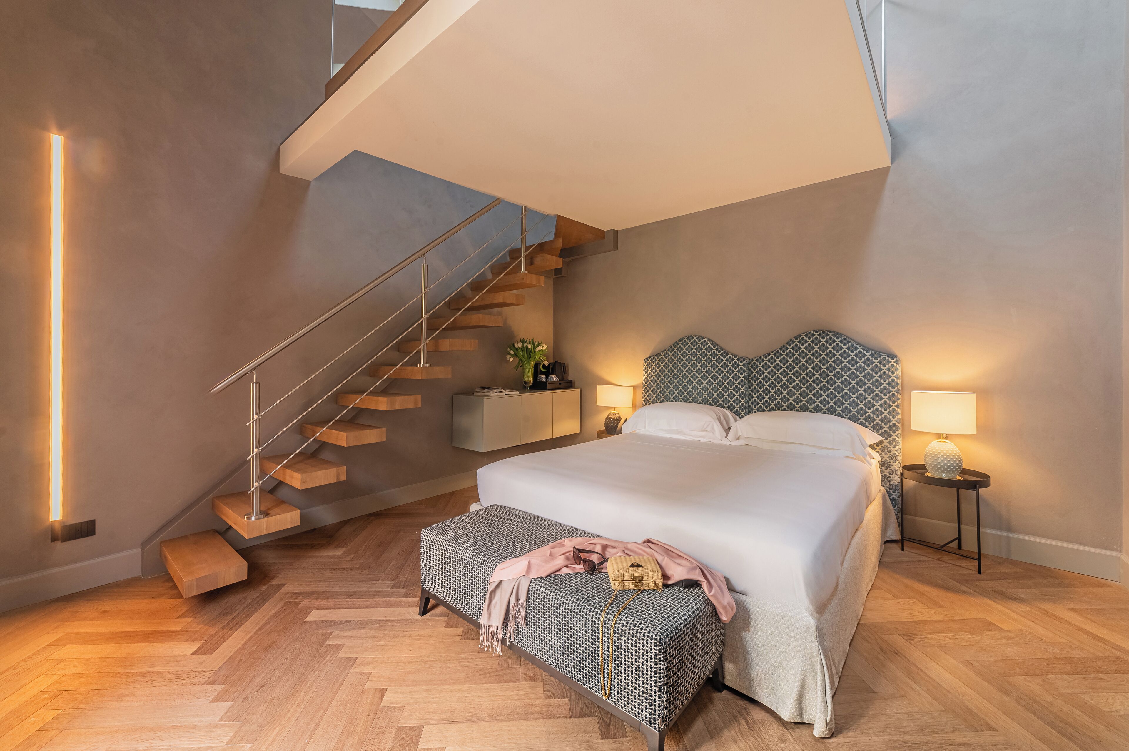 A modern bedroom with a large bed, a wooden staircase, and a nightstand with a lamp.
