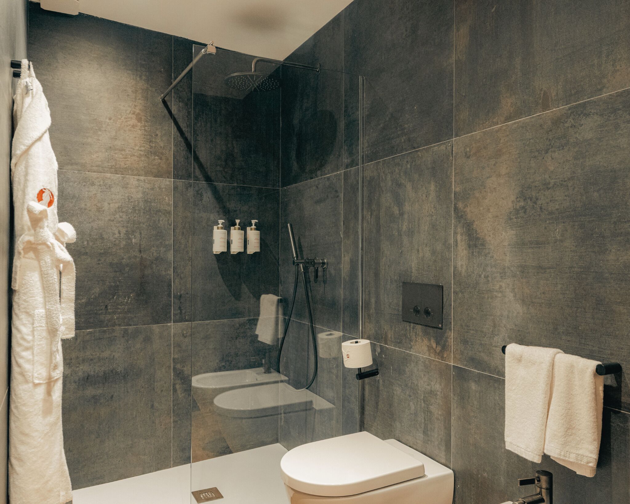 Une salle de bain moderne équipée d’une douche, de toilettes et d’un lavabo, avec des murs recouverts de carreaux foncés et des éléments en céramique blancs.