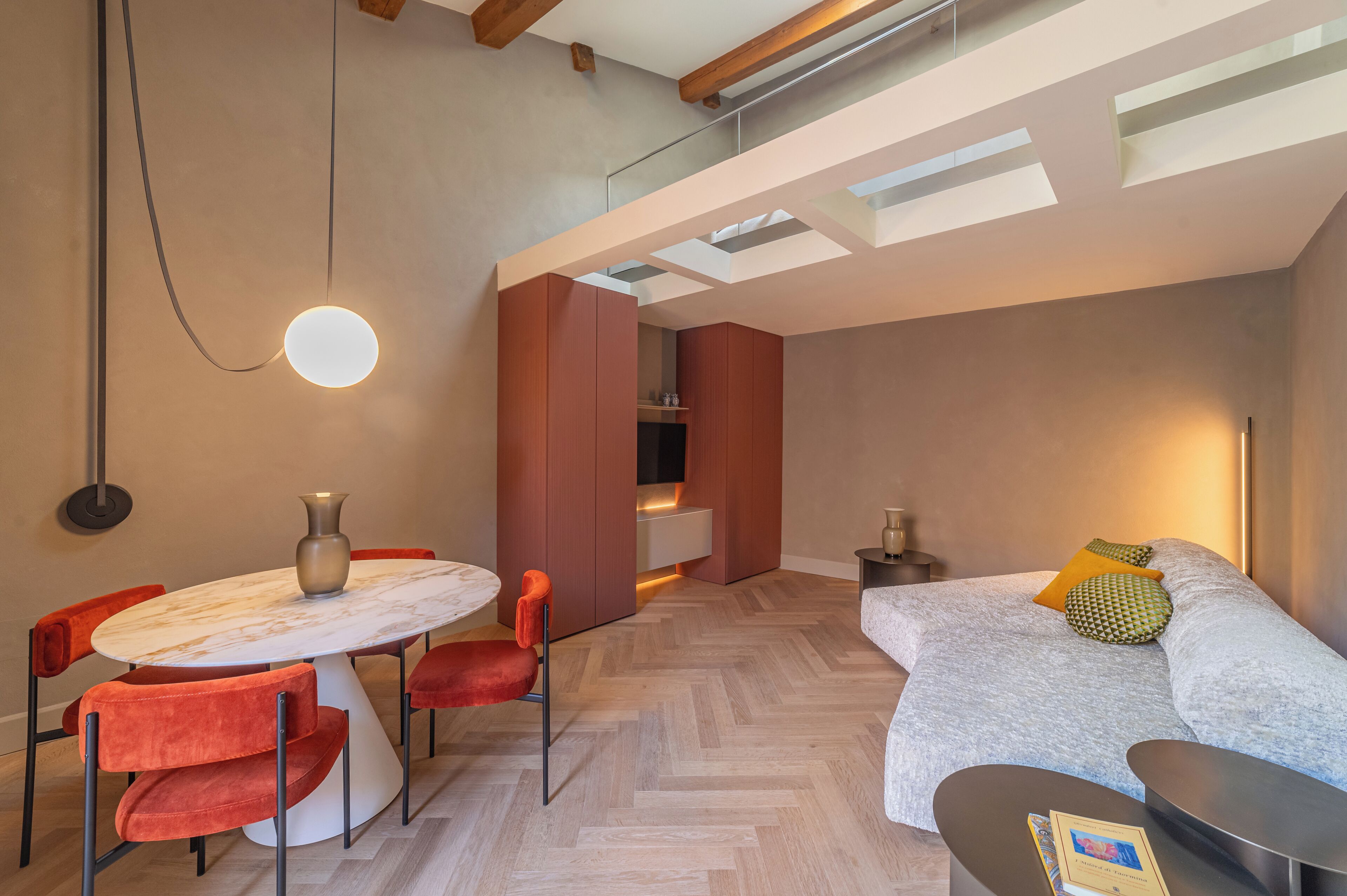 A modern, open-plan living and dining area with a herringbone wood floor, a round dining table, red upholstered chairs, and a sofa with a throw pillow.