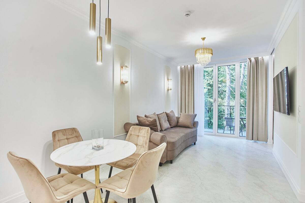 Ein modernes Wohnzimmer mit einem runden Esstisch, einem beigen Sofa und einem Balkon mit Blick ins Grüne.