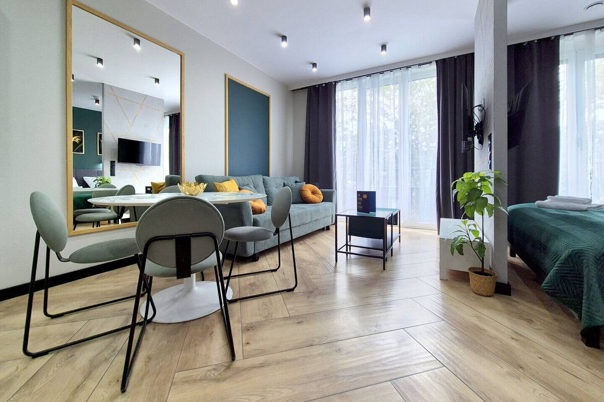 Ein modernes Hotelzimmer mit einer Couch, einem Esstisch mit Stühlen, einem kleinen Beistelltisch, einem Bett mit grüner Tagesdecke, einem großen Spiegel und einer Zimmerpflanze.