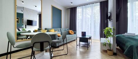 A modern hotel room with a sofa, a dining table with chairs, a small side table, a bed with a green bedspread, a large mirror, and a potted plant.