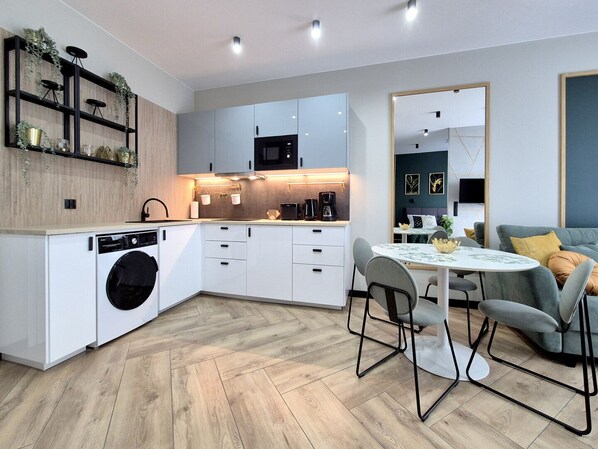 A modern kitchen with a built-in washing machine, a dining area with a round table and chairs, and a living area with a sofa and cushions.