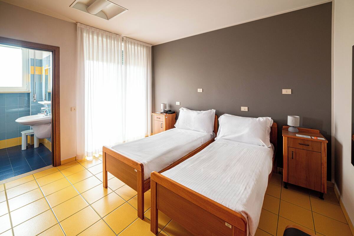 A hotel room with two single beds, a wooden nightstand, a bedside table, a small cabinet, and a bathroom visible through an open door.