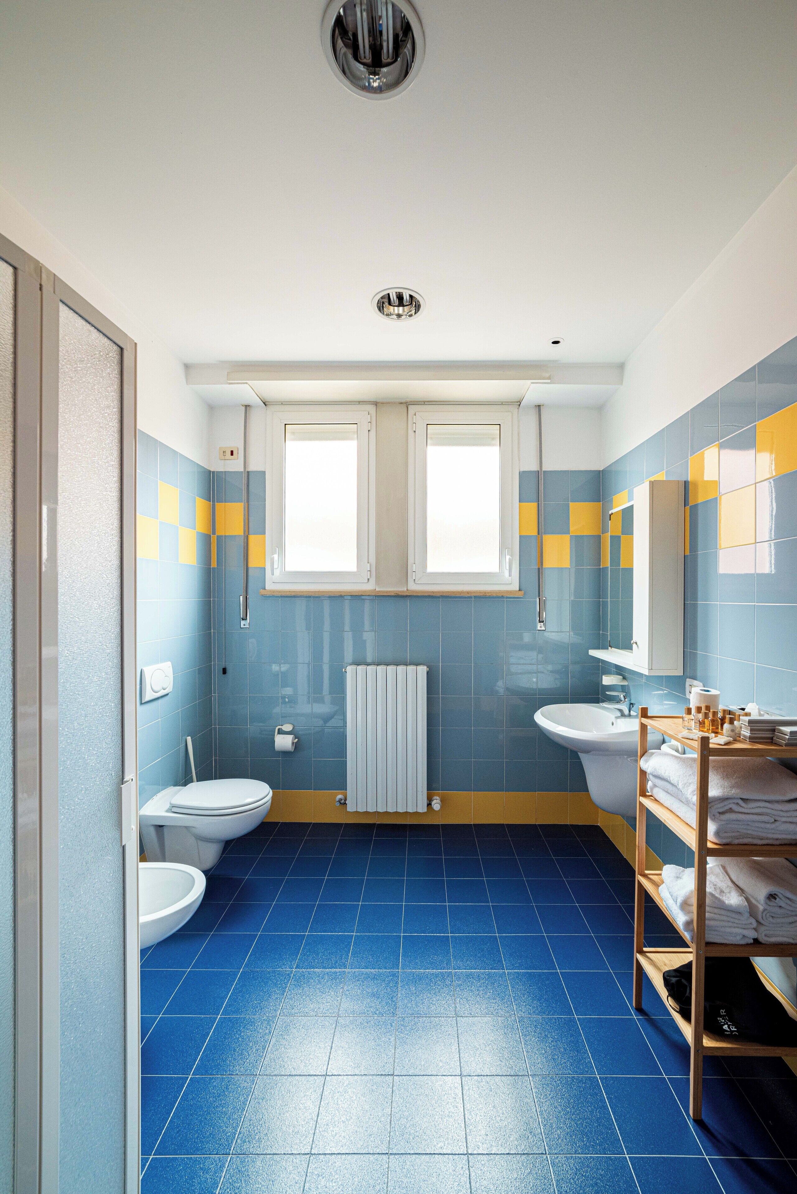 A bathroom with blue tiles, two toilets, a sink, a mirror, and a shelf with towels.