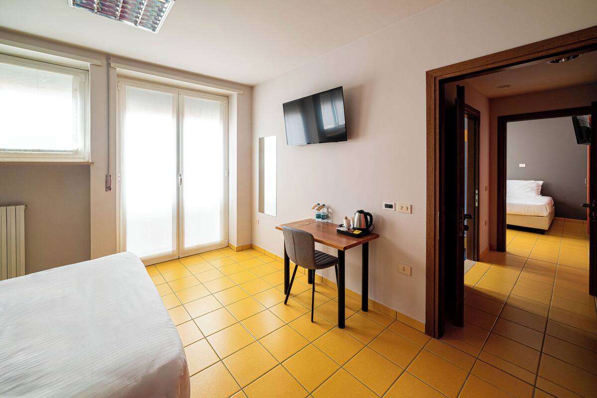 A hotel room with a bed, a desk with a chair, a flat-screen TV mounted on the wall, and a bathroom visible through an open door.