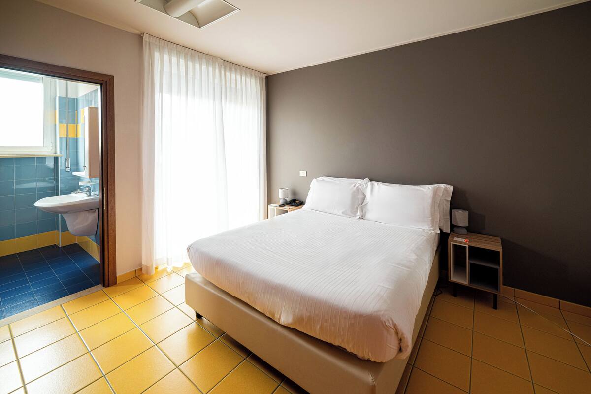 A hotel room with a bed, a nightstand, a bedside table, a window with curtains, and a bathroom visible through an open door.