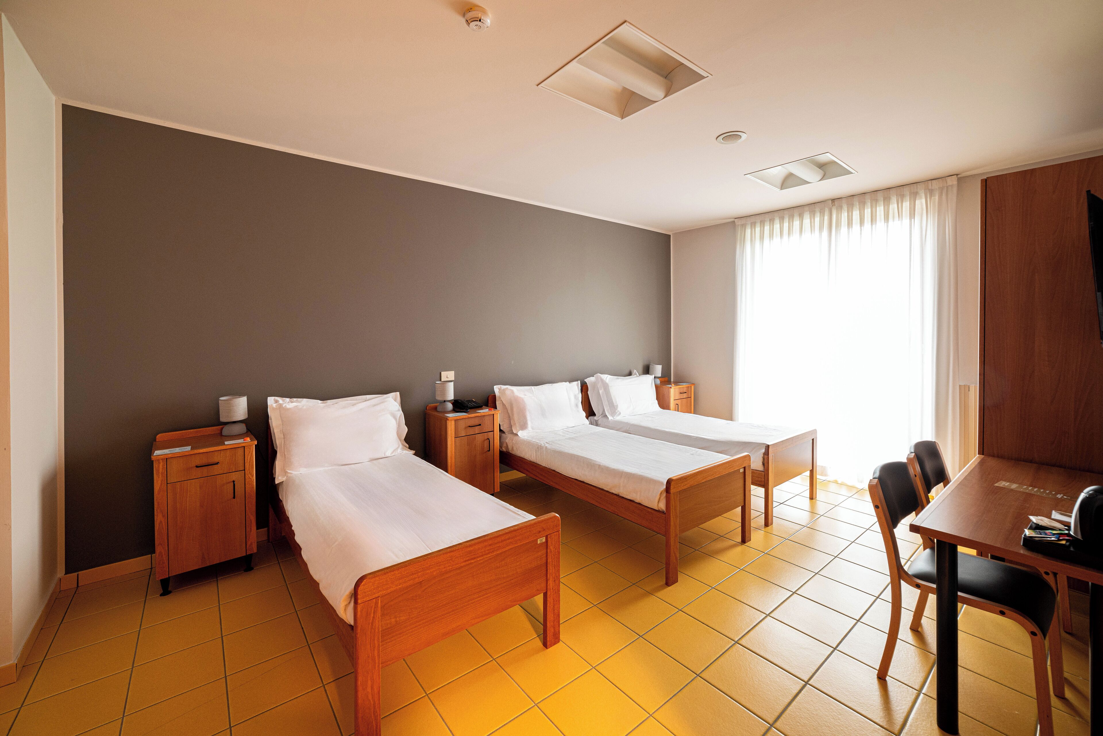 A hotel room with two single beds, a wooden nightstand with a lamp, a wooden table with chairs, and a large window with curtains.