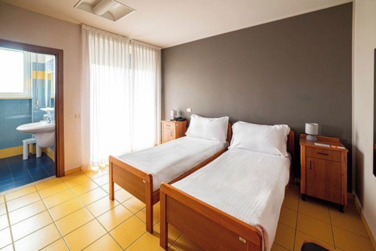 A hotel room with two single beds, a wooden nightstand, a small table, and a bathroom visible through an open door.