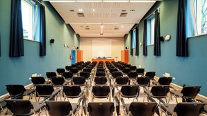 A conference room with rows of chairs facing a stage, blue walls, and large windows with curtains. - Phi Hotel Homy Druento (Druento)