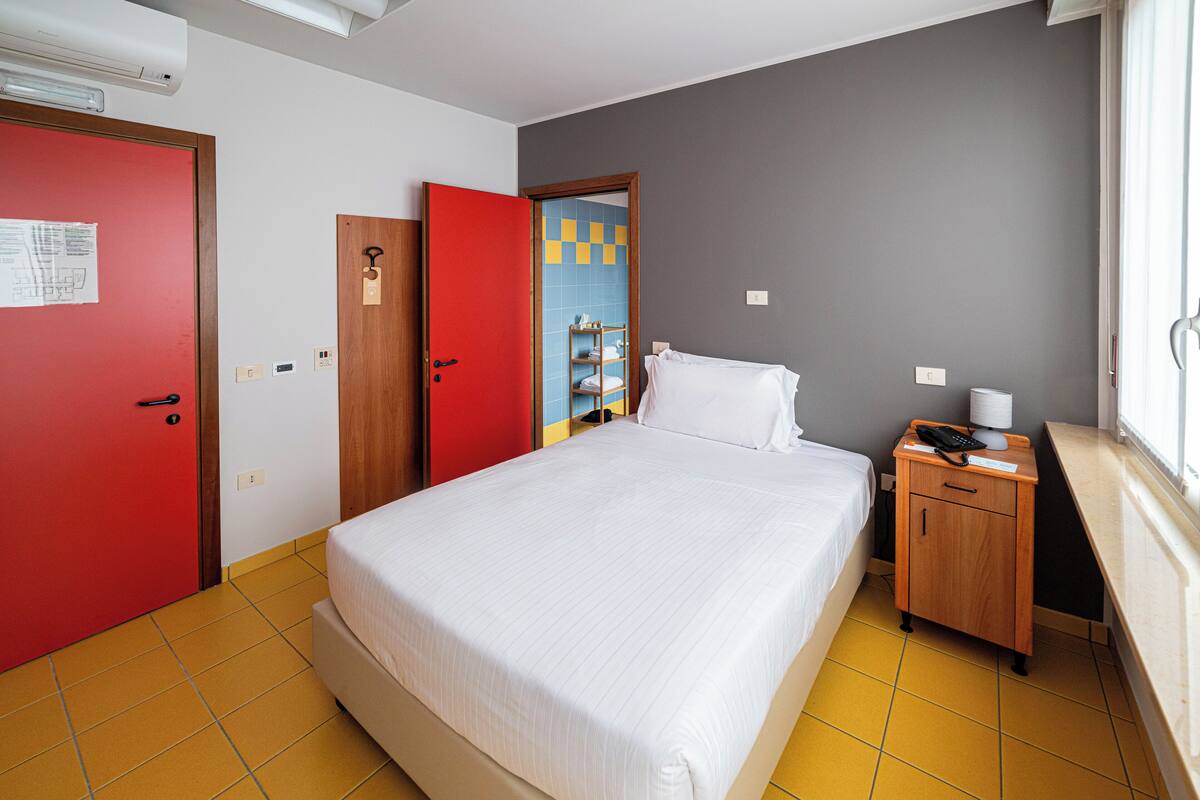 A hotel room with a bed, a desk with a lamp, a red door, and a bathroom visible through an open door.
