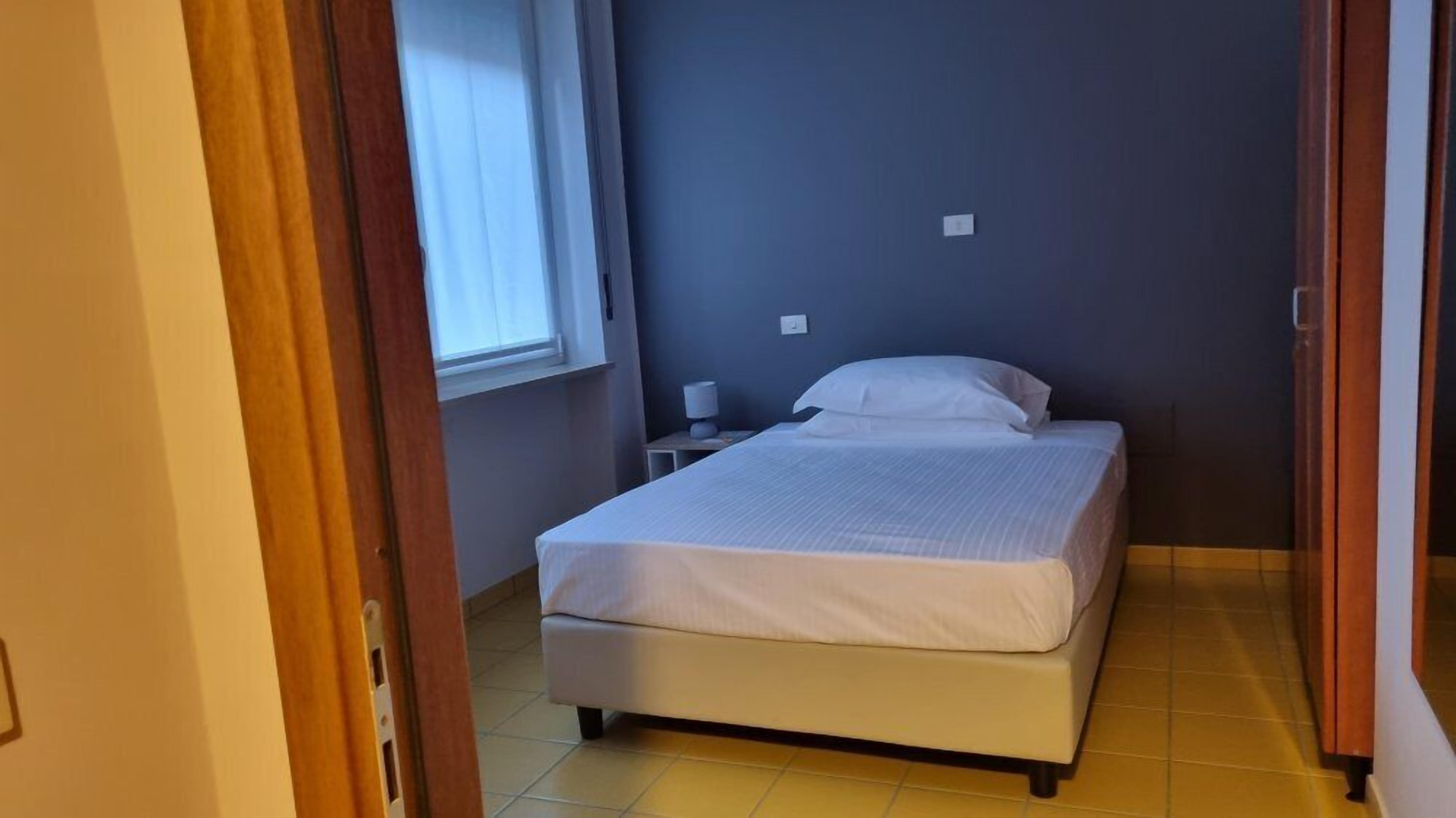 Una cama pequeña y bien hecha en una habitación con ventana, mesita de noche y toma de corriente.