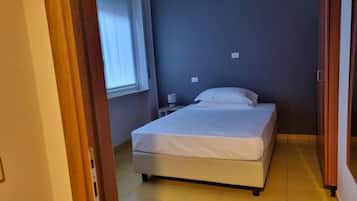 Una cama pequeña y bien hecha en una habitación con ventana, mesita de noche y toma de corriente.