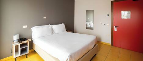 A hotel room with a bed, a nightstand, a red door, and a yellow-tiled floor.