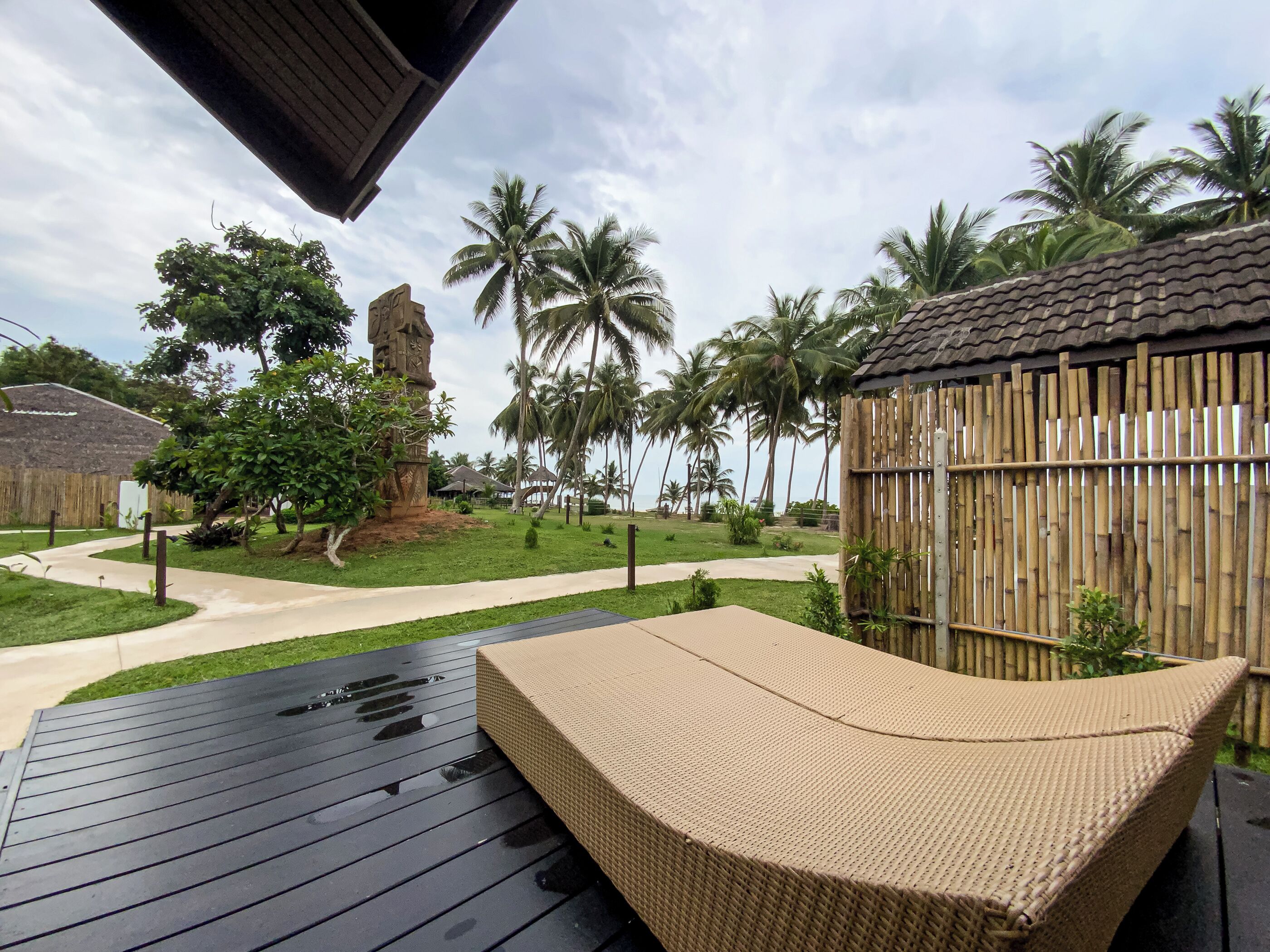 Una terraza de madera con una silla lounge tejida, un monumento de piedra y una valla de bambú.