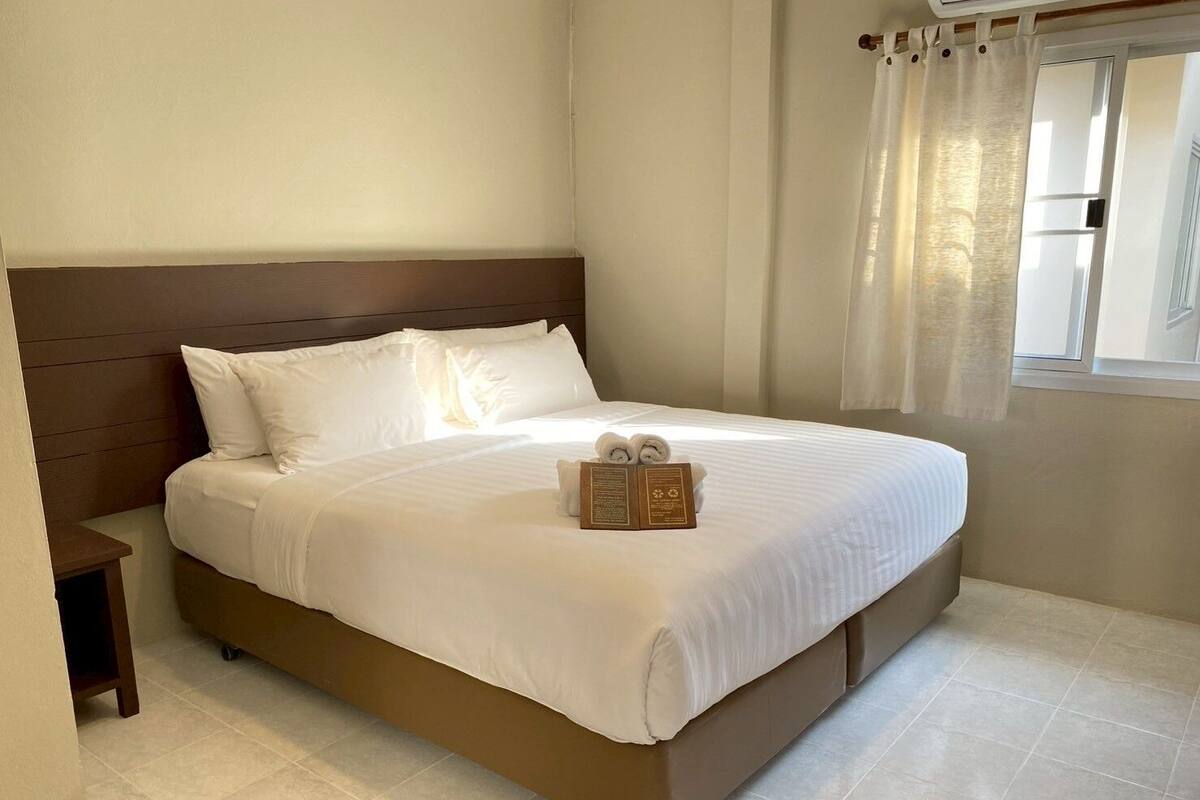 A neatly made bed with white linens and a wooden headboard, a bedside table, and a window with curtains.