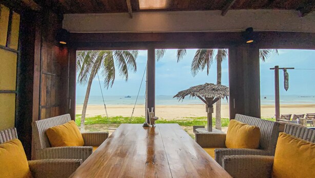 Una mesa de madera con sillas de mimbre y cojines amarillos, con vista a una playa con palmeras y una sombrilla de paja.