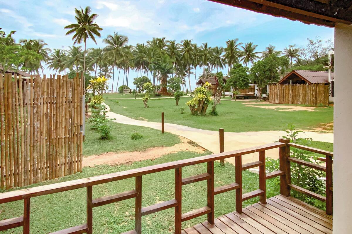 A wooden deck with a view of a well-maintained lawn, palm trees, and a small building in the distance.
