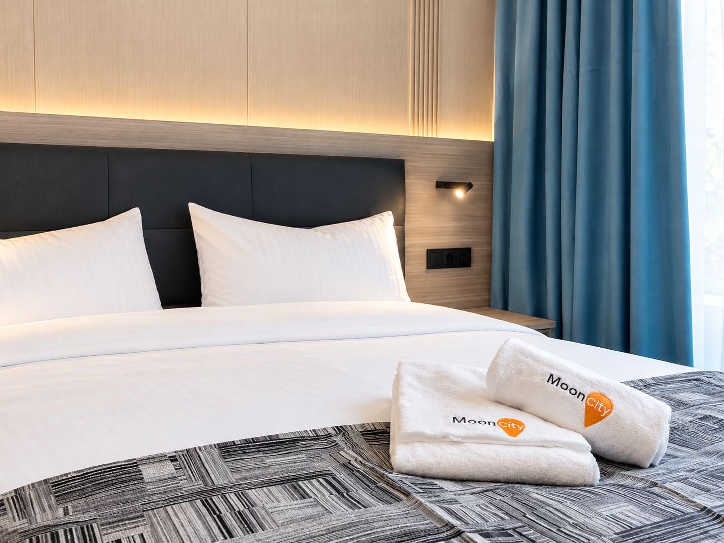 A hotel room with a bed, two pillows, a headboard, a nightstand, a lamp, and a window with curtains.