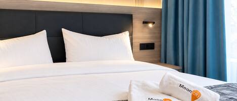 A hotel room with a bed, two pillows, a headboard, a nightstand, a lamp, and a window with curtains.