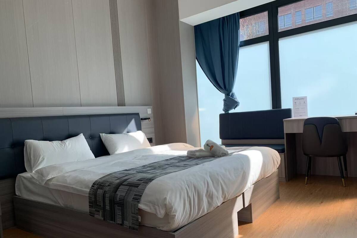 A modern hotel room with a large bed, a desk, and a view of the city through the window.