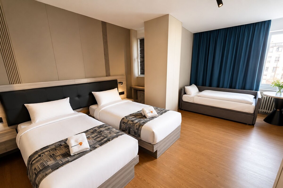 A hotel room with two beds, a sofa, a window with curtains, and a radiator.