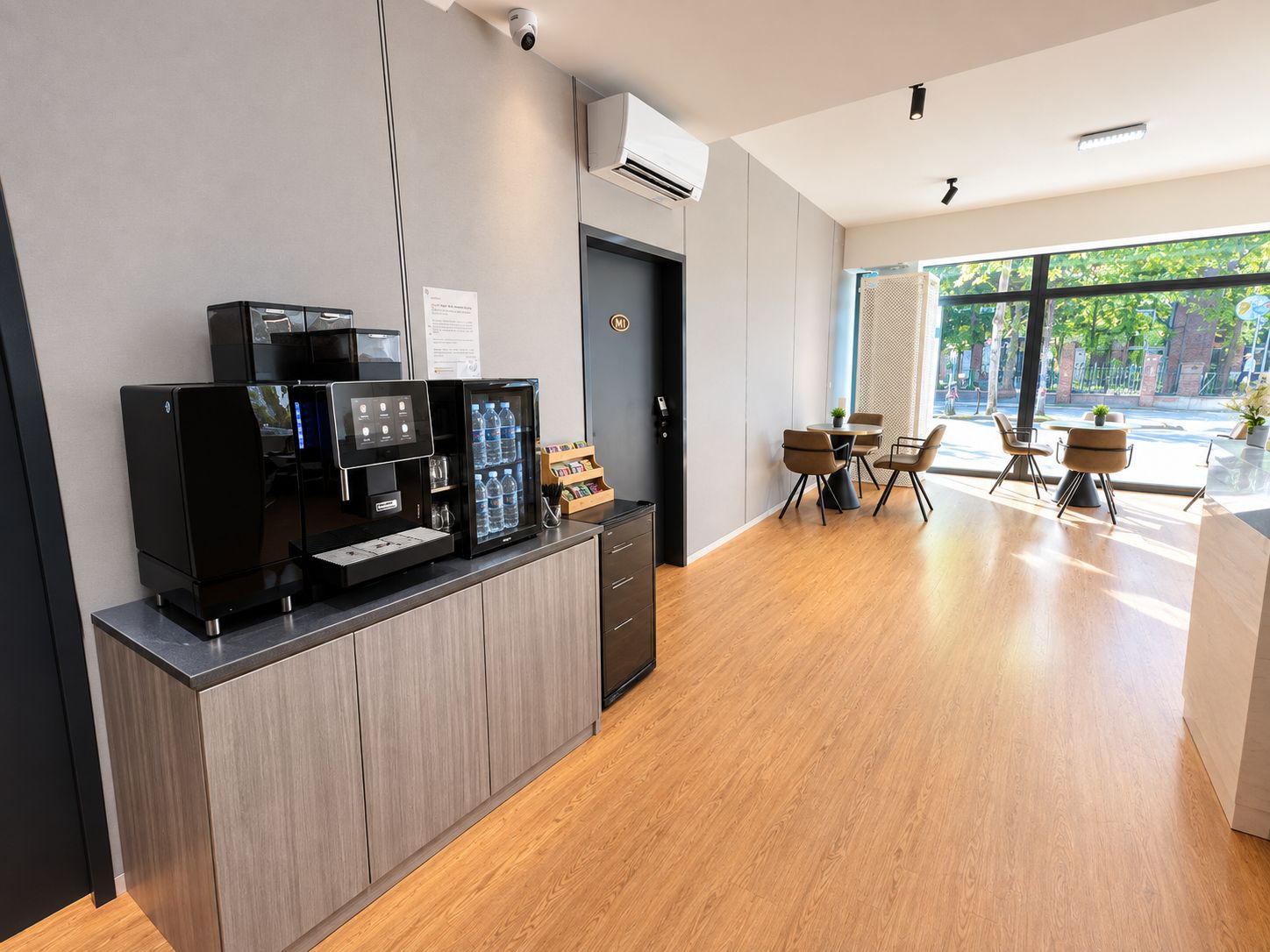 Ett modernt receptionsområde med en kaffemaskin, ett kylskåp och en sittgrupp med bord och stolar.