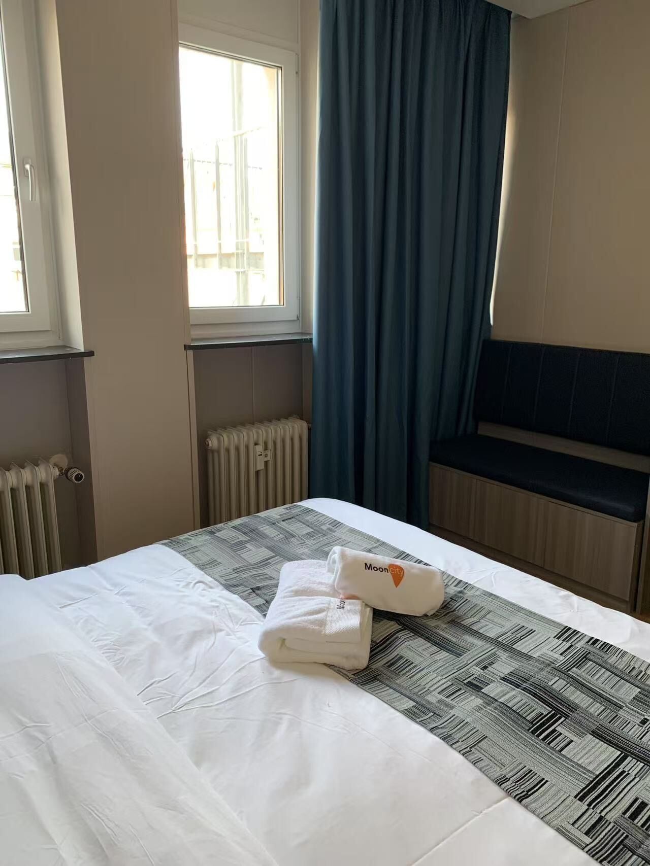 Una habitación de hotel con una cama, un ventanal con cortinas, un radiador y un banco.