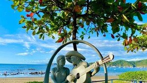 A statue of a man and woman sitting on a bench with a guitar, surrounded by greenery and overlooking a beach. - Coastal Comfort: Spacious Second-Floor Getaway (Playas Del Coco)