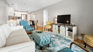 Un salon moderne avec un canapé, des fauteuils, un téléviseur et une cuisine en arrière-plan.