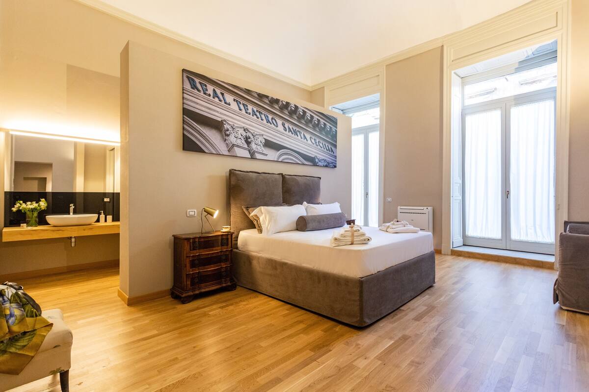 A modern bedroom with a large bed, a bedside table, a mirror, and a view of the outside.
