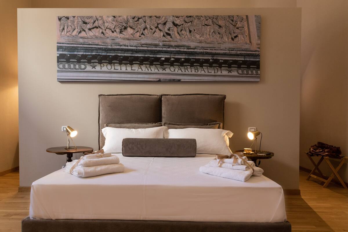 A neatly made bed with white linens and two pillows, a wall-mounted artwork depicting a historical scene, and two bedside tables with lamps.