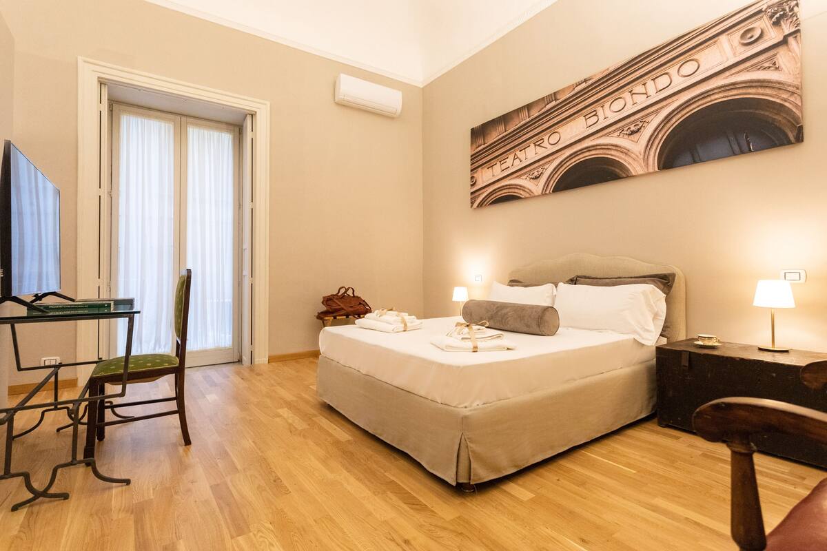 A hotel room with a bed, a desk, a chair, a TV, and a painting on the wall.