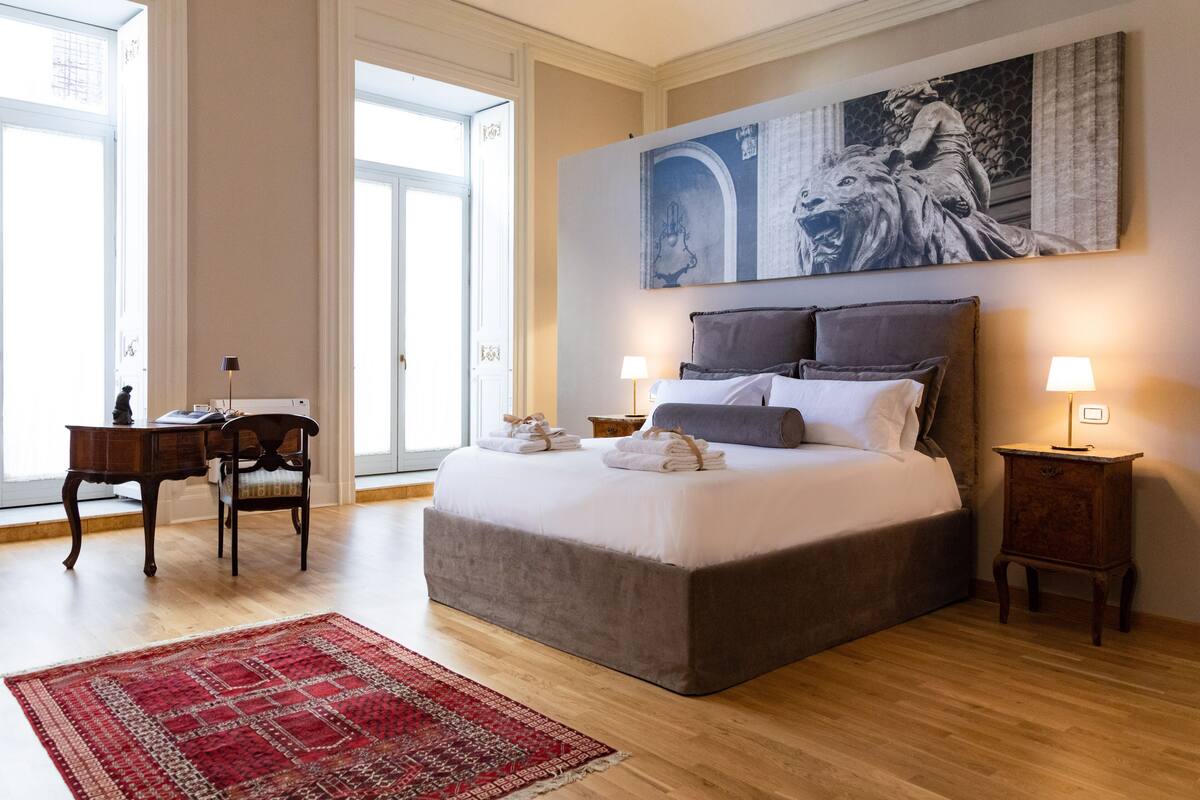 A bedroom with a large bed, a desk, a chair, a red rug, and a painting on the wall.