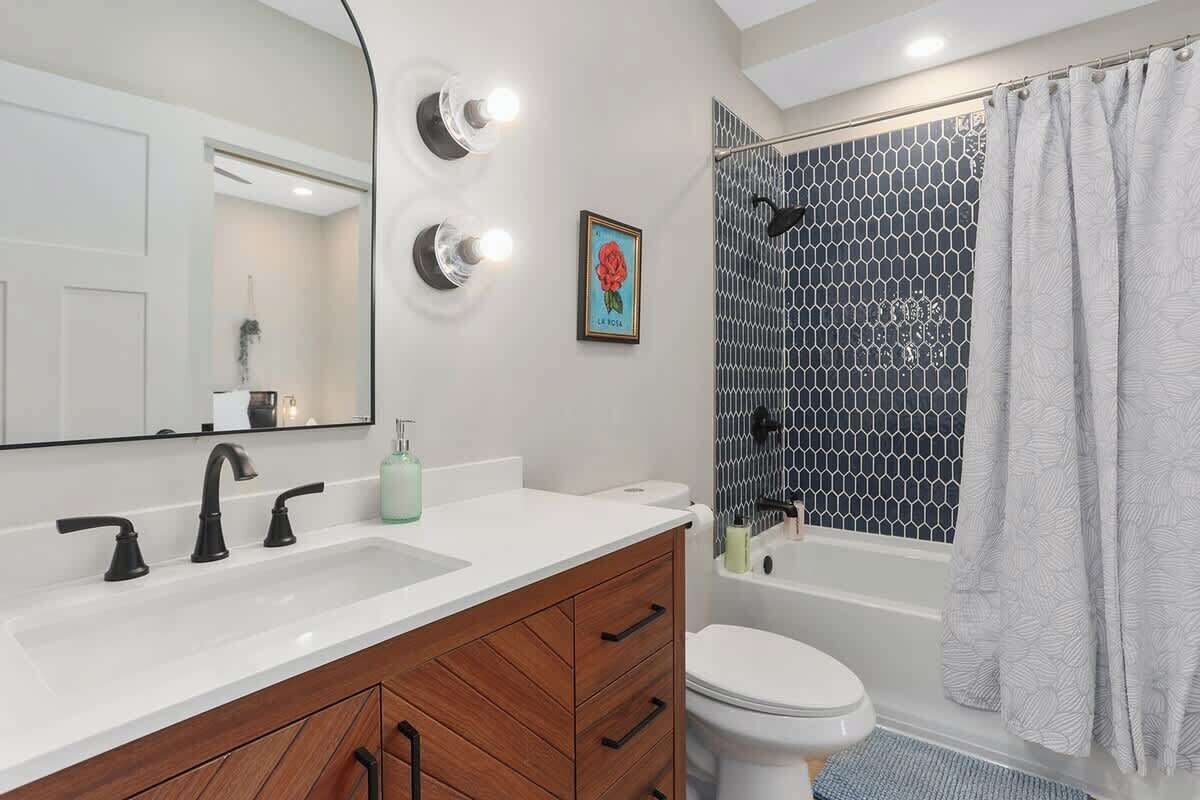 A bathroom with a white sink, black faucets, a wooden vanity, a white toilet, a shower with a black and white patterned wall, and a light-colored shower curtain.