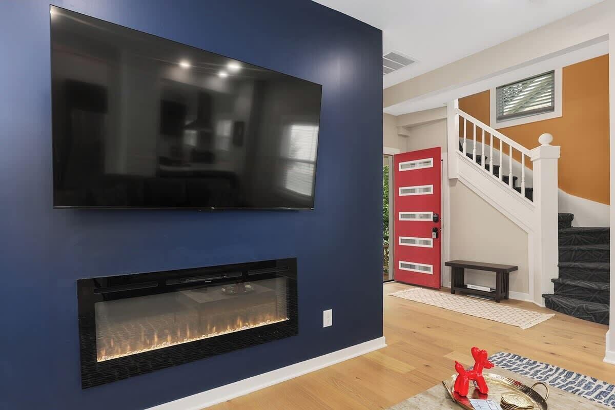 Un soggiorno moderno con una TV a schermo piatto montata su una parete blu scuro, un caminetto sottostante, una porta rossa e una scala con ringhiere bianche.