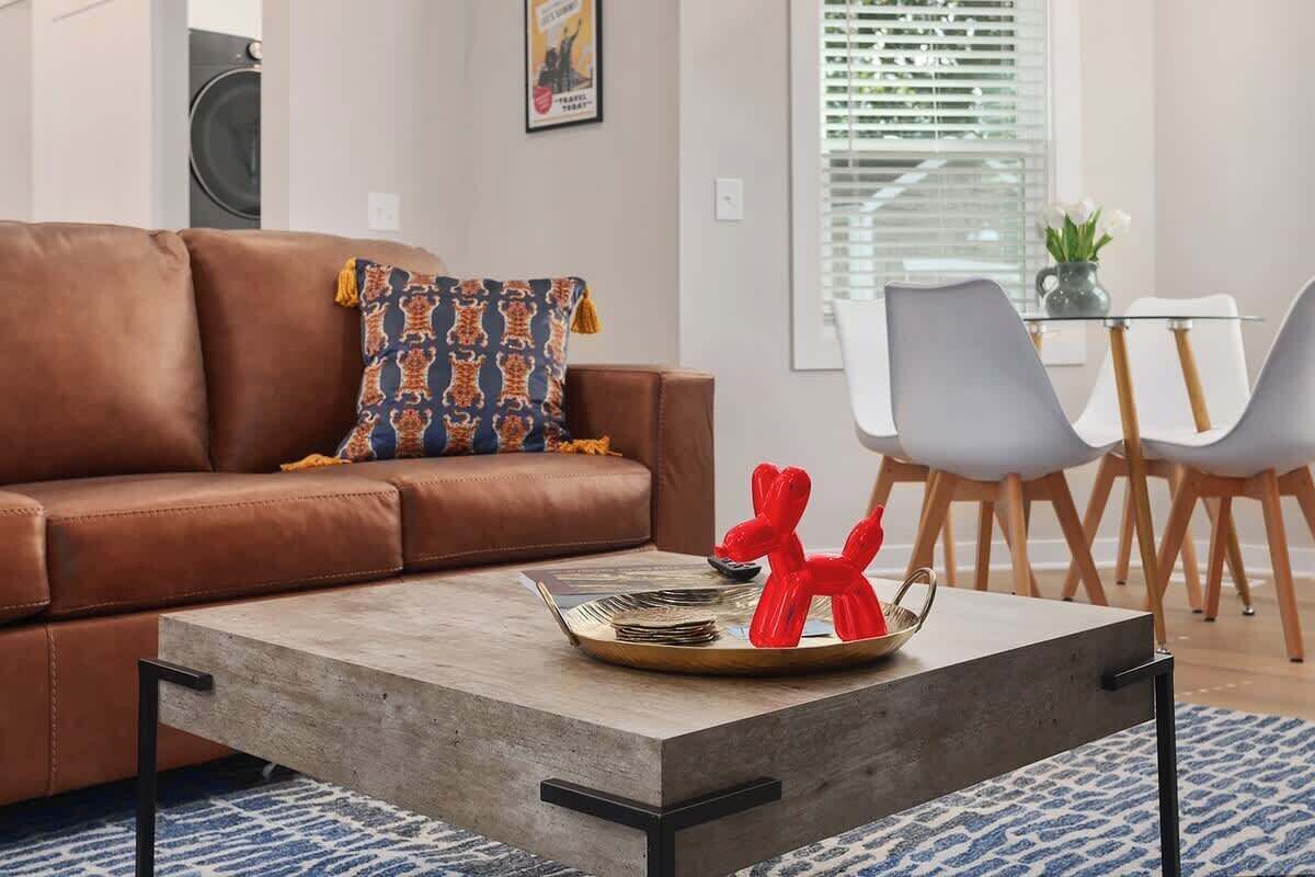 Un salon avec un canapé marron, un coussin décoratif à motifs, une table basse sur laquelle se trouve un plateau rempli de pièces de monnaie et une sculpture rouge, ainsi qu’un coin repas avec une table en verre et des chaises grises.