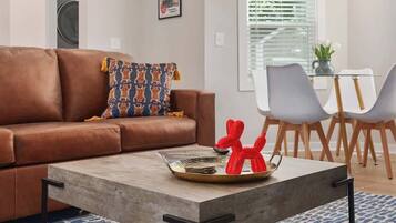 Un salon avec un canapé marron, un coussin décoratif à motifs, une table basse sur laquelle se trouve un plateau rempli de pièces de monnaie et une sculpture rouge, ainsi qu’un coin repas avec une table en verre et des chaises grises.
