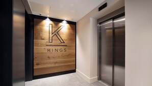 Een moderne lift met een metalen afwerking, naast een wand met een houten paneel waarop het logo en de naam "KINGS" staan.