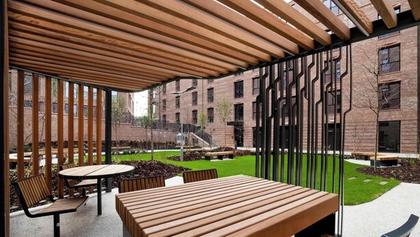 Een terras buiten met houten tafels en banken, een bakstenen gebouw op de achtergrond, en een gazon.