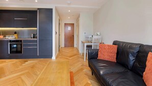 Een moderne keuken met donkere kastjes, een houten visgraatvloer, een zwarte leren bank met oranje kussens, en een eethoek met een witte tafel en stoelen.