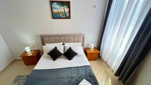 A neatly made bed with a headboard, two bedside tables with lamps, a painting on the wall, and a window with curtains.