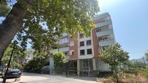 A multi-story building with a ground-level entrance, a parking area, and a tree-lined street.
