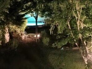 Un espacio exterior nocturno con piscina, rodeado de árboles y un patio con sillas y una mesa.