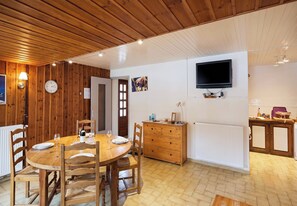 Een eetruimte met een houten tafel en stoelen, een klok, een aan de muur bevestigde televisie en een deur naar een andere kamer.
