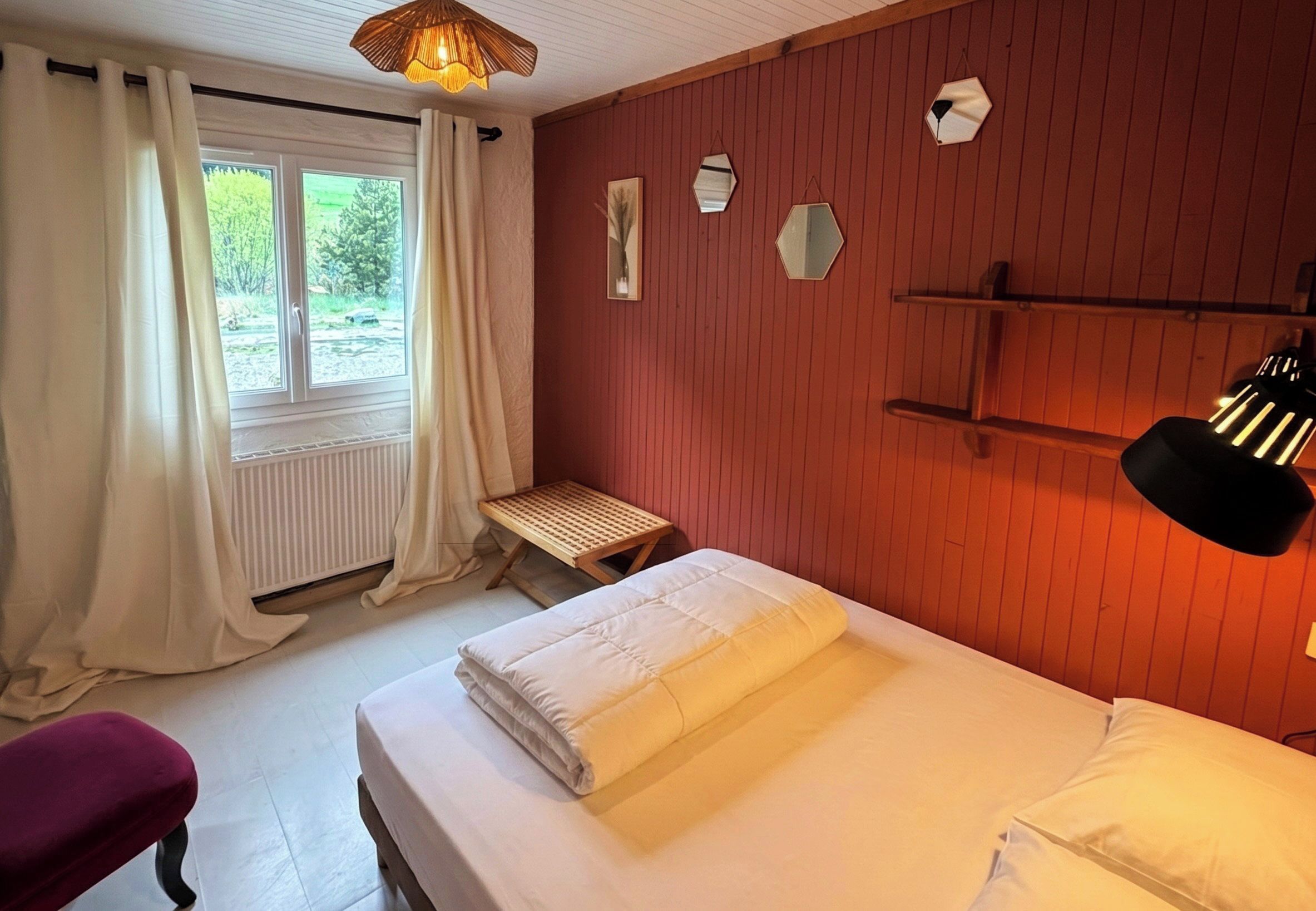 A bedroom with a bed, a bench, a window with curtains, and a wall-mounted shelf.