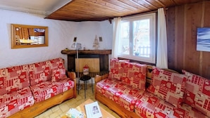 Een gezellige kamer met twee houten banken, een kleine tafel, een open haard en een raam met gordijnen.